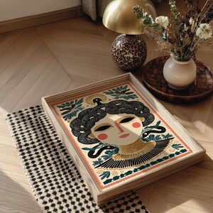 May include: Wooden serving tray featuring a stylized illustration of a woman with snakes in her hair. The artwork uses a palette of cream, black, gold, and teal. The tray has a light wood frame and is placed on a patterned rug.