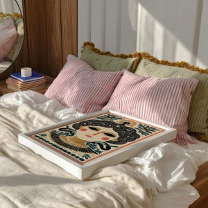 May include: A white framed art print featuring a stylized illustration of a woman's face with black hair and a gold headdress. The artwork is set on a bed with pink striped pillows and green pillows with yellow trim.