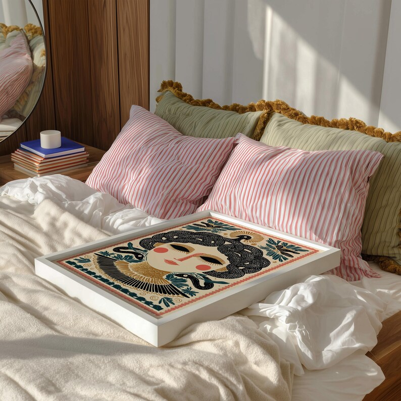 May include: A white framed art print featuring a stylized illustration of a woman's face with black hair and a gold headdress. The artwork is set on a bed with pink striped pillows and green pillows with yellow trim.