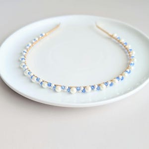 May include: A delicate headband featuring a gold-tone metal band adorned with alternating white faux pearls and small blue beads. The decorative elements are evenly spaced around the band, creating a refined and elegant accessory. The headband is displayed on a white plate.