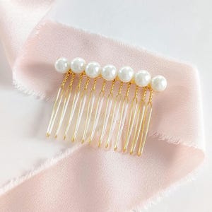 May include: A gold-toned hair comb featuring a row of seven round white faux pearls attached to the top edge. The comb has ten thin, parallel metal prongs extending downward, each connected to the pearl base with a delicate twisted gold wire.