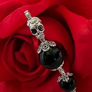 May include: A silver-toned skull-shaped decorative pin with black gemstone eyes and two black gemstone beads. The pin is set against a blurred red background, suggesting a floral theme.