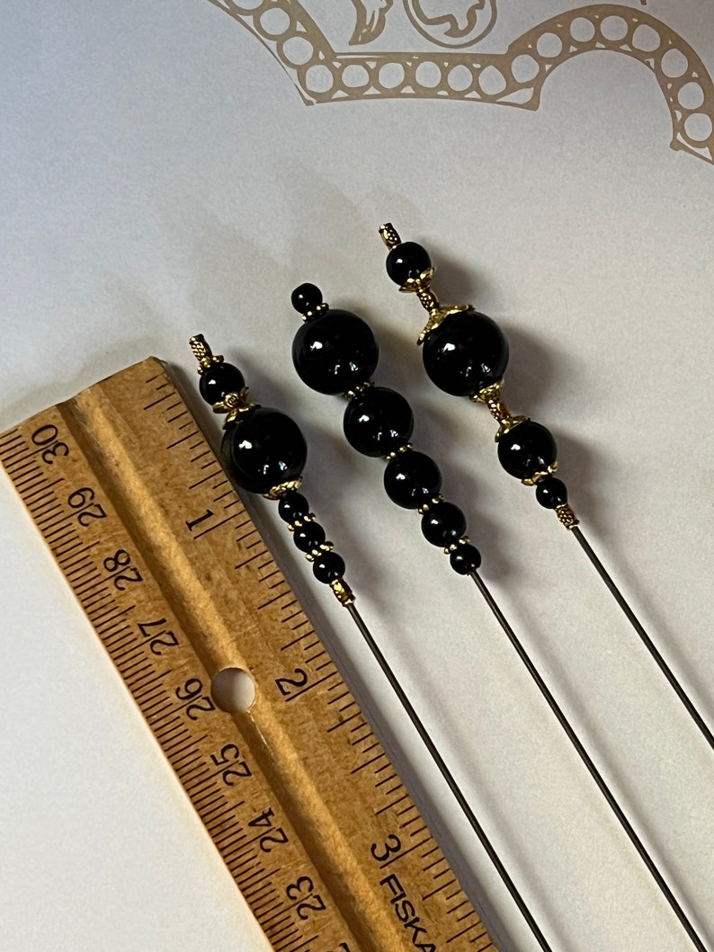 May include: Three black beaded hair pins with gold-colored accents. The pins feature a series of black beads of varying sizes, with gold-colored metal detailing. A wooden ruler is in the image, showing the pins' length.