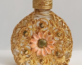 Op de afbeelding: Een sierlijke, vintage parfumfles. De heldere glazen fles is omhuld met een gouden filigraan ontwerp. Een roze en groene bloem is gecentreerd op de voorkant. De fles heeft een gouden, gedetailleerde dop.