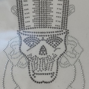 May include: A silver and black skull with a top hat made of rhinestones. The skull is surrounded by two silver ribbons.