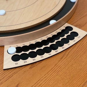 May include: A wooden cribbage board with black scoring pegs and white playing pieces. The board has a circular playing area with concentric circles and a scoring track with numbers from 0 to 100.