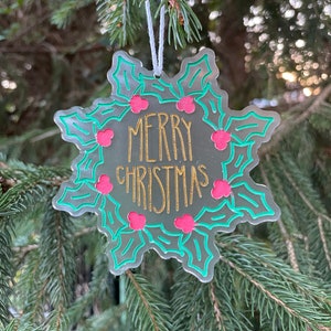 May include: A silver snowflake-shaped Christmas ornament with a green holly leaf design and red berries. The ornament says "Merry Christmas" in gold lettering.