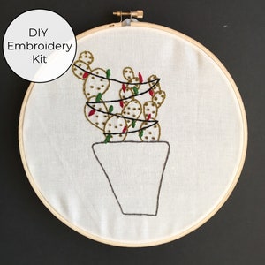 May include: DIY Embroidery Kit featuring a cactus with green and red embroidery floss in a white pot. The embroidery is on white fabric in a wooden embroidery hoop.