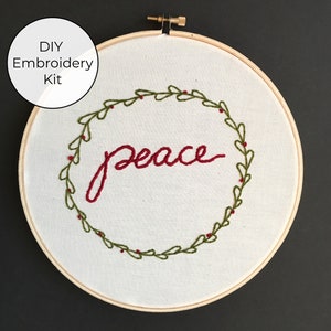 May include: DIY embroidery kit featuring a white hoop with a green wreath and red berries surrounding the word "peace" in red embroidery thread.