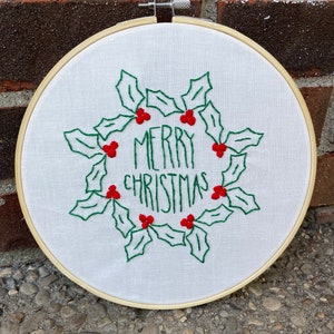 May include: Embroidered hoop art with a white background and green holly leaves with red berries. The text "Merry Christmas" is embroidered in green thread.