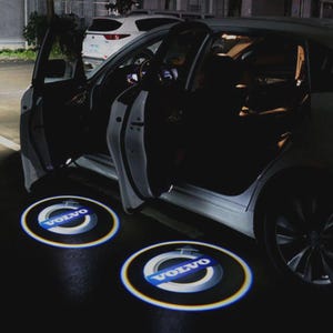 Può includere: Un'auto Volvo bianca con le portiere aperte, che proietta loghi illuminati sul terreno. I loghi sono circolari, con la scritta "VOLVO" in bianco e blu. La scena è illuminata dai fari dell'auto di notte.