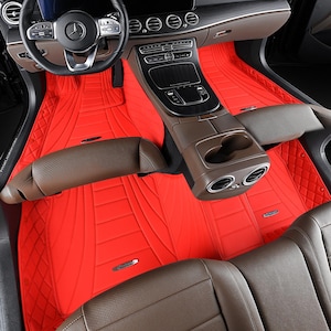 Puede incluir: Alfombrillas rojas brillantes para coche con un patrón de diamantes acolchados y detalles de costura. Las alfombrillas están hechas a medida para el interior de un vehículo, cubriendo los huecos para los pies delanteros y traseros. El interior también presenta una consola central y asientos marrones.