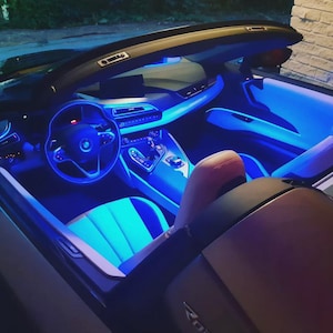 Puede incluir: Vista interior de un coche descapotable con iluminación ambiental azul. Se ven el salpicadero, el volante y los asientos, destacando el diseño moderno y las características iluminadas del coche. Los asientos son una combinación de azul claro y negro.