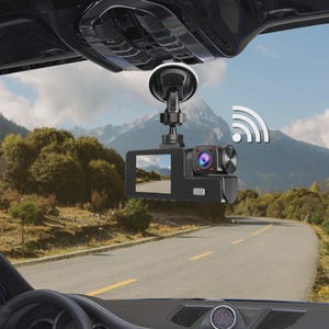 Può includere: Dash cam nera montata sul parabrezza di un'auto, che mostra una scena stradale. La telecamera ha uno schermo, un obiettivo e un simbolo Wi-Fi. Lo sfondo mostra una strada che si snoda attraverso un paesaggio montuoso.