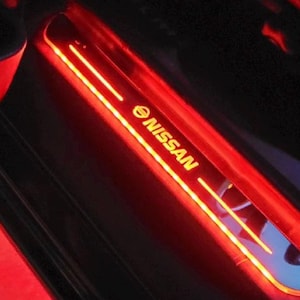 May include: Illuminated red and yellow Nissan door sill plate. The plate features the Nissan logo and brand name in yellow, with two parallel yellow light strips. The surrounding area is black.