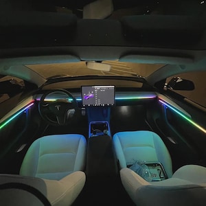 May include: Interior view of a Tesla car with ambient lighting. The car's dashboard features a large touchscreen display and a steering wheel with the Tesla logo. The seats are white, and the car is illuminated with colorful LED lights.