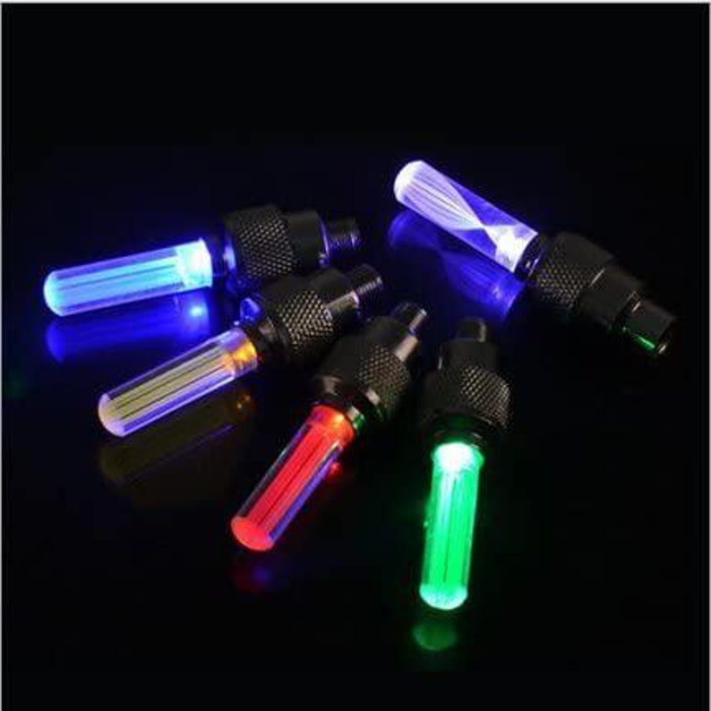 May include: A collection of black tire valve stem lights with glowing LED bulbs in blue, red, and green. The lights have a cylindrical shape and are arranged against a dark background.