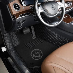 May include: Black leather car floor mats with a diamond pattern. The mats are installed in a luxury car with brown leather seats. The mats have a logo with the letter "M" in the centre.