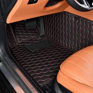 May include: Black leather car floor mats with red stitching and a honeycomb pattern. The mats are installed in a car with tan leather seats.