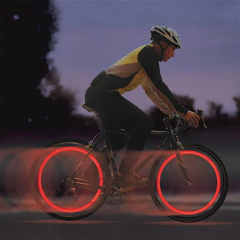 May include: A person riding a bicycle at night with illuminated red lights on the wheels. The bicycle is black, and the person is wearing a helmet and dark clothing. The lights create a glowing effect.