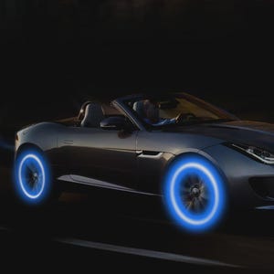 May include: A silver convertible car with glowing blue wheel lights driving on a dark road. The car's wheels are illuminated with a bright blue light, creating a futuristic effect. The driver is visible inside the car.