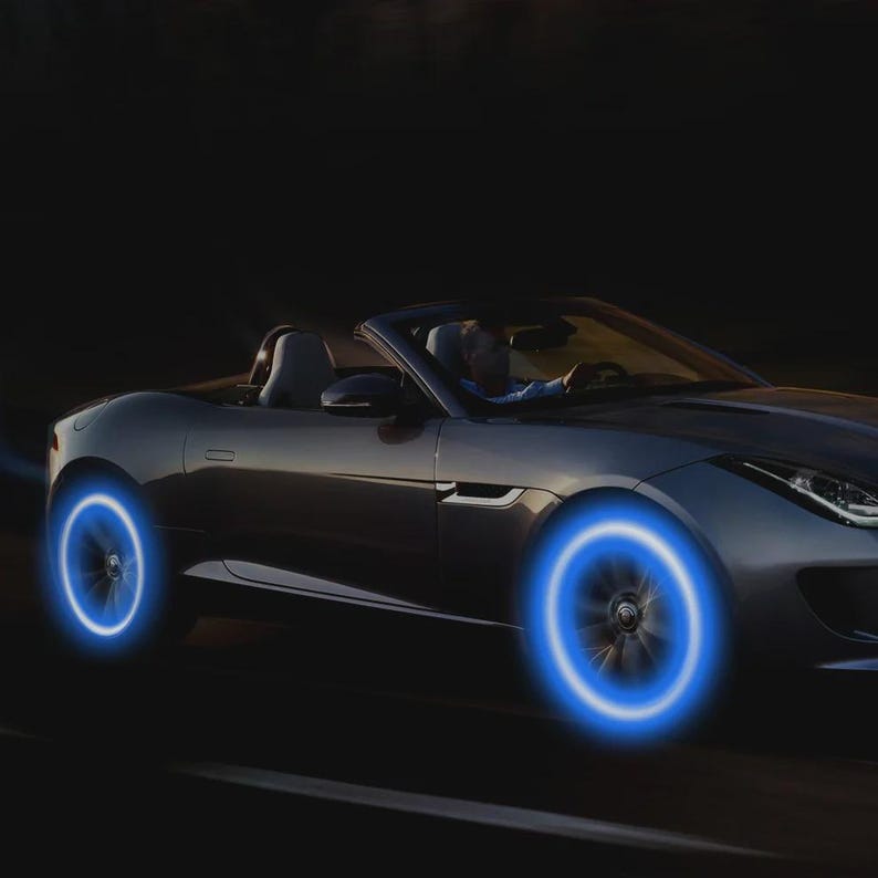 May include: A silver convertible car with glowing blue wheel lights driving on a dark road. The car's wheels are illuminated with a bright blue light, creating a futuristic effect. The driver is visible inside the car.
