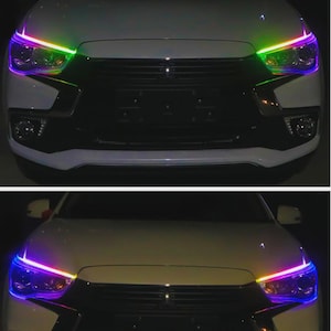 Peut inclure: Avant d'une voiture blanche avec des phares éclairés. Les lumières affichent un dégradé de couleurs, notamment le violet, le vert et le jaune. Le pare-chocs et la calandre de la voiture sont visibles.
