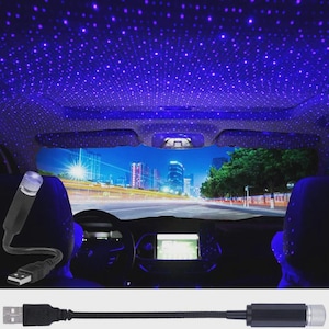 Könnte beinhalten: Ein Auto-Innenraum mit Sternenhimmel-Effekt an der Decke, erzeugt durch einen USB-betriebenen LED-Projektor. Das Bild zeigt das Armaturenbrett, die Sitze und eine Skyline durch die Windschutzscheibe. Der Projektor ist schwarz mit silbernem Oberteil.
