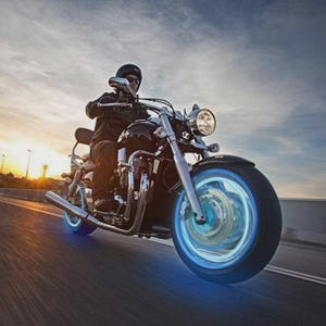 May include: A black motorcycle with blue glowing wheels is shown in motion on a road. The rider is wearing a helmet and dark clothing. The sky is a mix of blue and orange, suggesting a sunset or sunrise.