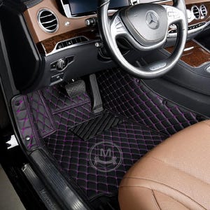 May include: Black car floor mats with a diamond pattern and purple stitching. The mats are installed in a luxury car with brown leather seats.