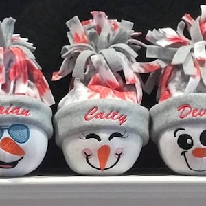 May include: Five gray and white snowman ornaments with red and white pom-poms on top. Each snowman has a different embroidered face and a gray hat with a name embroidered on it: John, Maureen, Brian, Caitly, and Devon.