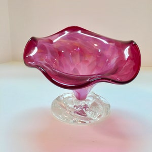 May include: A pink glass vase with a clear glass base. The vase has a ruffled, flower-like shape.