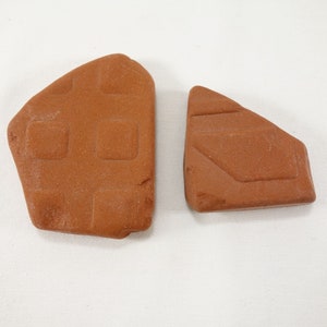 Puede incluir: Dos baldosas de cerámica de color marrón rojizo de forma irregular. Una baldosa presenta un patrón de rejilla de cuadrados empotrados, mientras que la otra tiene un diseño rectangular empotrado con líneas anguladas. Las baldosas están sobre un fondo blanco.