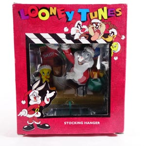 May include: A Looney Tunes stocking hanger featuring Bugs Bunny dressed as Santa Claus, Tweety Bird, and other characters. The box is red with the Looney Tunes logo and character illustrations. The words "STOCKING HANGER" are printed on the box.