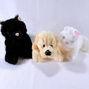 May include: Three plush toys consisting of a black cat with a pink nose, a light tan dog with a black nose, and a white cat with a pink nose and a small pink bow on its head. Each animal has soft, textured fur and is positioned in a row.