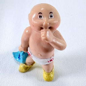 Puede incluir: Una figura vintage de Cabbage Patch Kids de un bebé con la cabeza calva, que lleva un pañal con detalles rojos y zapatos amarillos. El bebé sostiene un objeto azul y tiene el dedo en la boca.