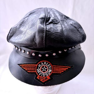 May include: A black leather cap featuring a textured crown and a smooth brim. A black leather band with silver-toned metal studs encircles the base. An embroidered patch displays the text Harley Davidson USA with orange wings and a central star.