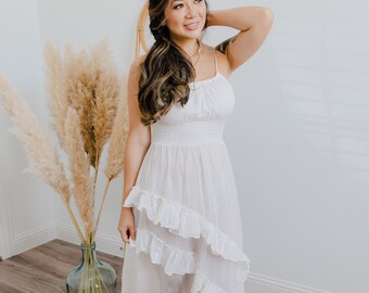 May include: A woman wearing a white, ruffled midi dress with a scoop neck and spaghetti straps. She is standing on a wooden floor with a vase of dried pampas grass behind her.