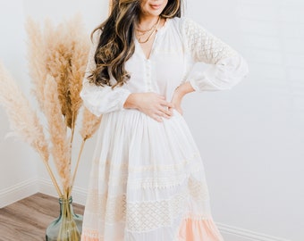 May include: A white midi dress with a lace overlay and a peach-coloured ruffle hem. The dress has long sleeves and a button-up front. The model is wearing a gold necklace.