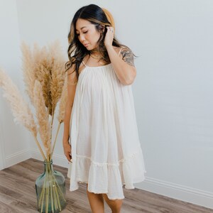 May include: A woman wearing a white, sleeveless, tiered dress with a ruffled hem. She is standing on a wooden floor with a vase of dried pampas grass behind her.