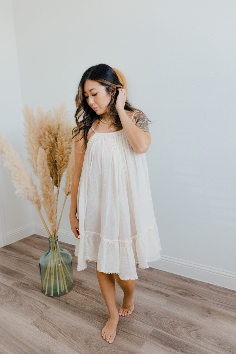 May include: A woman wearing a white, sleeveless, tiered dress with a ruffled hem. She is standing on a wooden floor with a vase of dried pampas grass behind her.