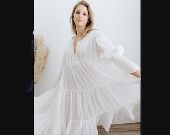May include: A white, oversized dress with a pin tuck detail and a V-neckline. The dress has a tiered skirt and long sleeves.