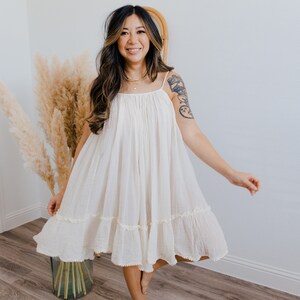 May include: A woman wearing a white, tiered, sleeveless dress with a ruffled hem. She is standing on a wooden floor with a vase of dried pampas grass behind her.