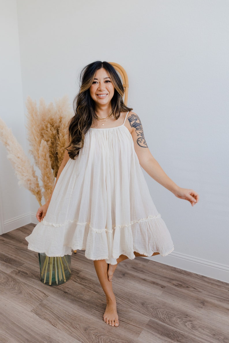 May include: A woman wearing a white, tiered, sleeveless dress with a ruffled hem. She is standing on a wooden floor with a vase of dried pampas grass behind her.