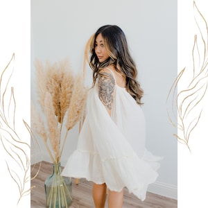 May include: A woman wearing a white, off-the-shoulder dress with a ruffled hem. She has long, dark hair and a tattoo on her arm. The dress is flowing in the wind.