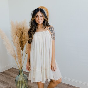 May include: A woman wearing a white, sleeveless, cotton dress with a ruffled hem. The dress has a scoop neck and a relaxed fit. She is standing in front of a white wall with a vase of dried pampas grass.