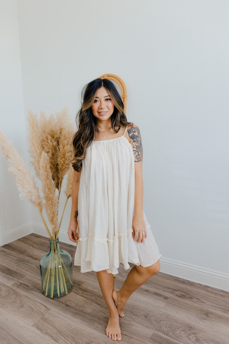 May include: A woman wearing a white, sleeveless, cotton dress with a ruffled hem. The dress has a scoop neck and a relaxed fit. She is standing in front of a white wall with a vase of dried pampas grass.