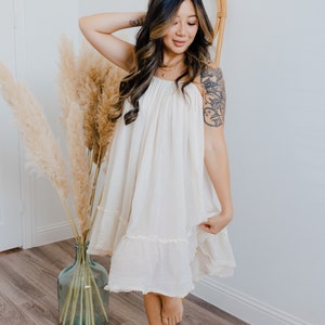 May include: A woman wearing a white, flowing dress with a ruffled hem. She is standing in front of a white wall with a vase of dried pampas grass.