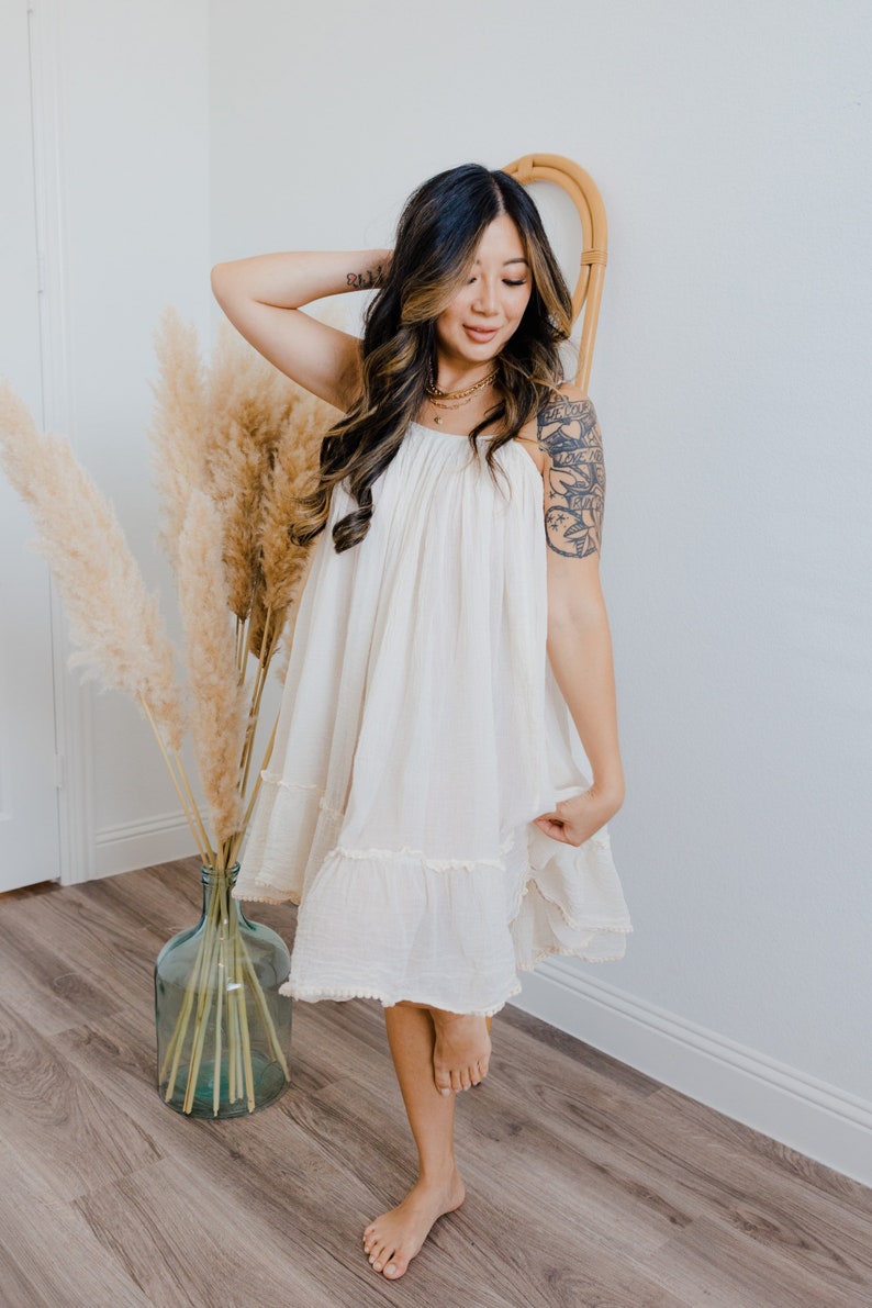 May include: A woman wearing a white, flowing dress with a ruffled hem. She is standing in front of a white wall with a vase of dried pampas grass.