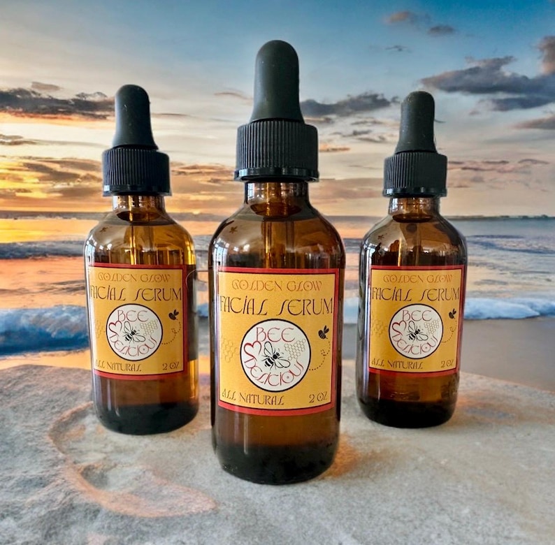 May include: Three amber glass bottles of Golden Glow Facial Serum with black droppers. Each bottle has a yellow label with the text "BEE DELICIOUS" and "ALL NATURAL". The bottles are 2 oz each.